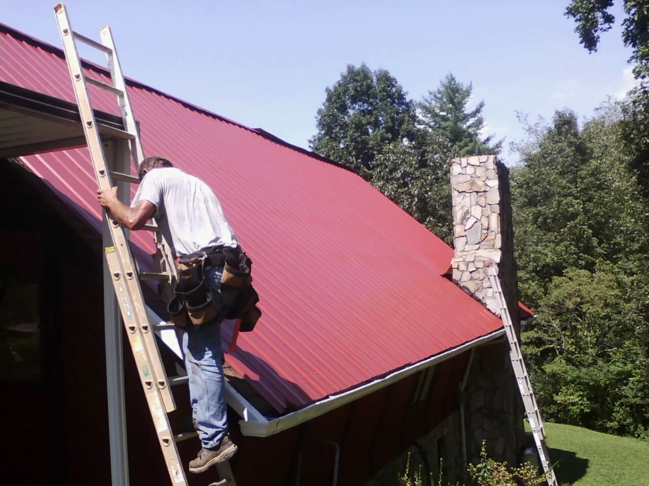 Roofer on a ladder beside a red metal roof and stone chimney — Soffit & Fascia Repair by Roofing Brothers in Tenmile